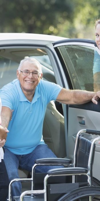 A senior man smiling at the camera as he goes from back seat of car into a wheelchair. His wife is holding his hand and his adult daughter is behind the wheelchair, holding the car door open.